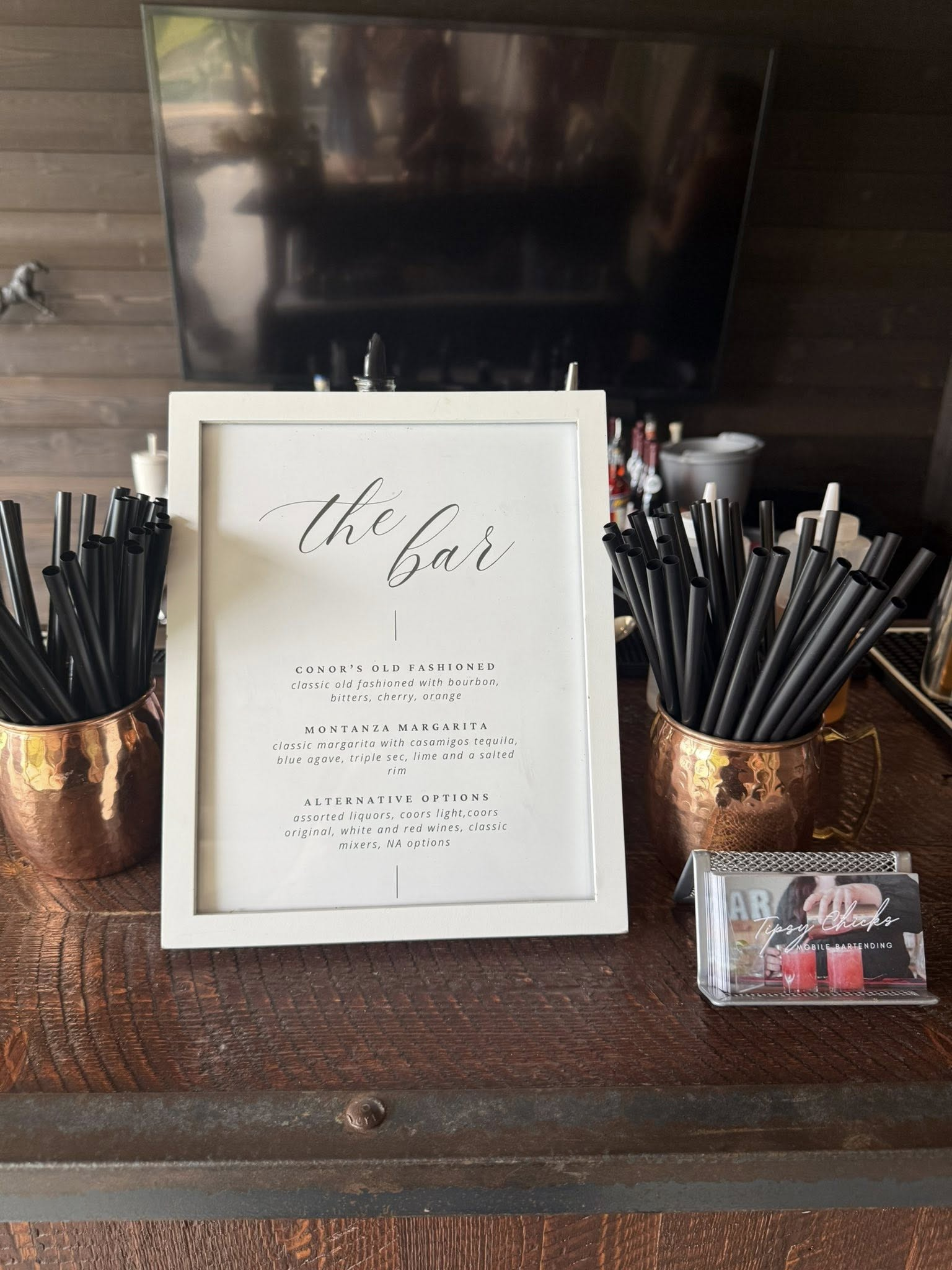 A white framed menu titled 'the bar' on a wooden bar countertop, with black straws in copper-colored holders on either side, and a small clear business card holder with promotional cards to the right.