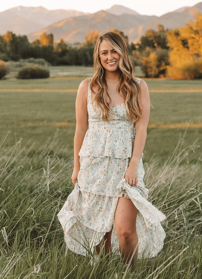 A young woman with long wavy blonde hair smiling in a grassy field with mountains and trees in the background during sunset.