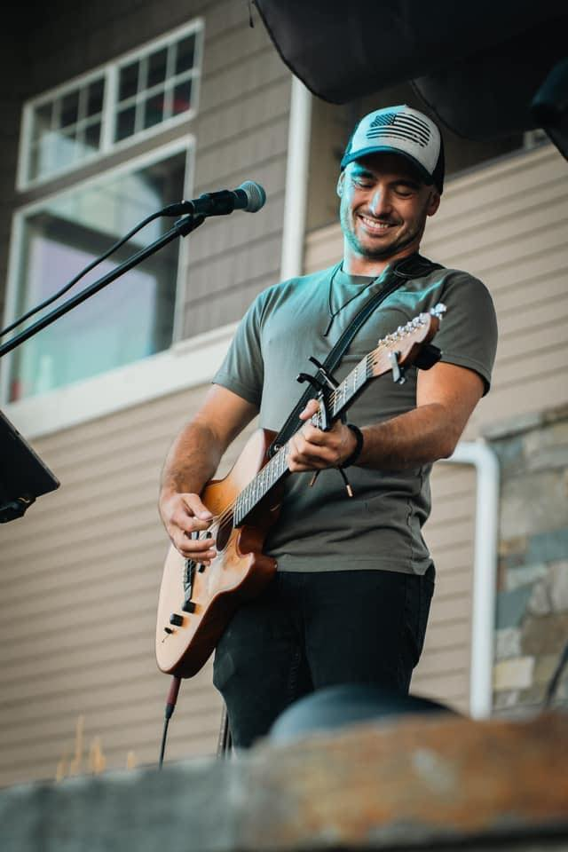 A man smiling while playing an acoustic guitar with a microphone and speaker nearby, outdoors at a house with large windows and beige siding.