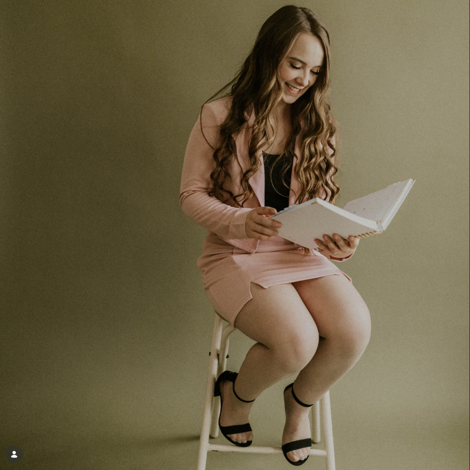 A woman with long, wavy brown hair sitting on a stool, smiling while reading a large white book. She's wearing a pink blazer over a black top, a pink skirt, and black sandals with ankle straps, against a plain, muted background.