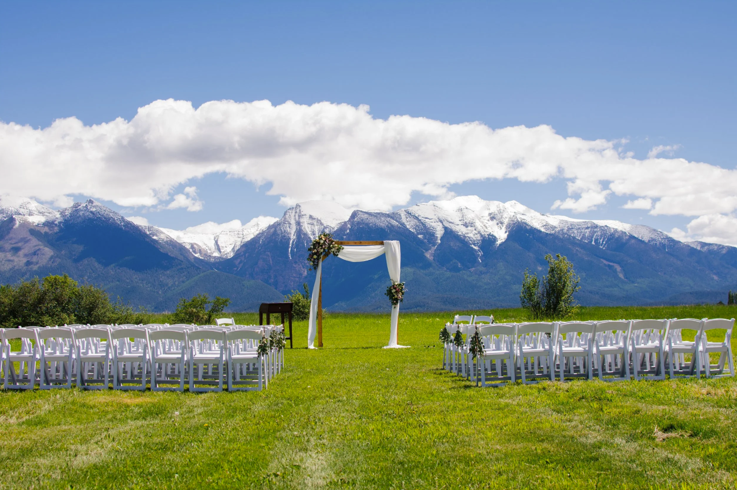 Outdoor wedding ceremony setup on a grassy field with white chairs, an arch decorated with flowers and drapery, and snowy mountain range in the background under a partly cloudy sky.