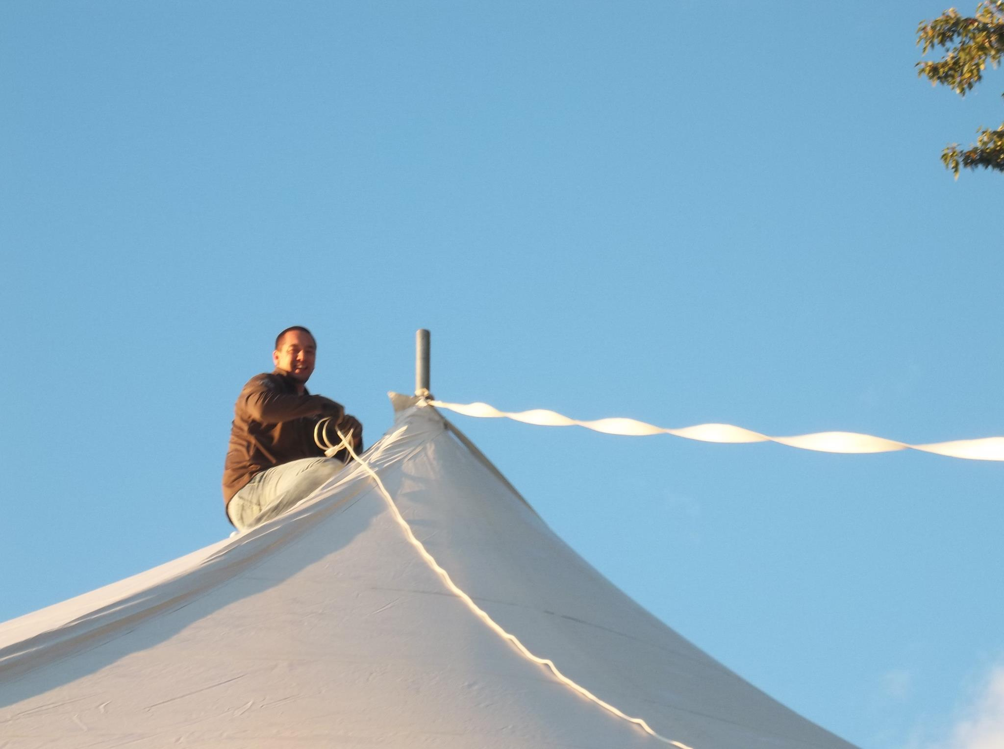 A person holding a flag or banner on top of a white, cone-shaped structure against a clear blue sky, with some tree leaves visible in the top right corner.