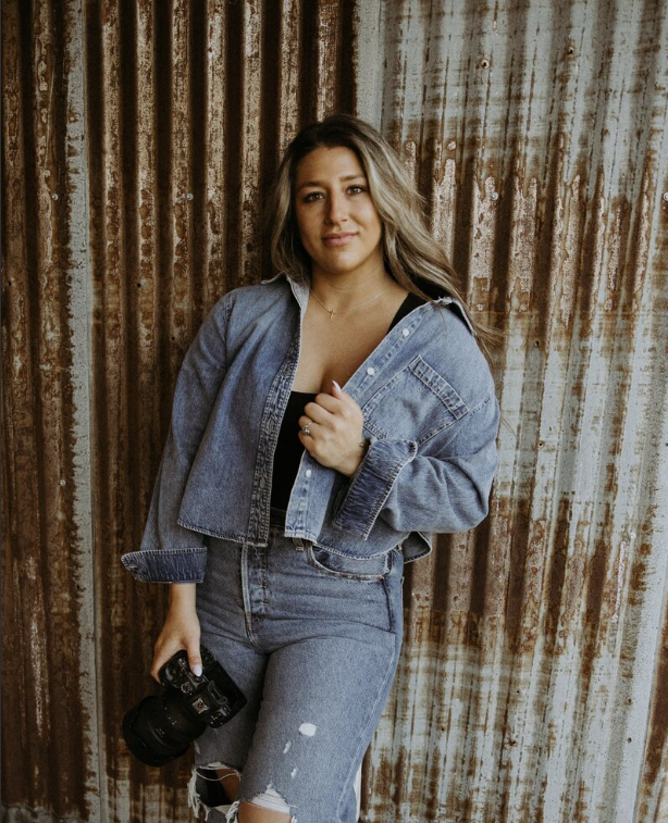 Young woman with long wavy hair holding a camera, standing in front of a rusted metal wall, wearing a denim jacket over a black top and ripped jeans.