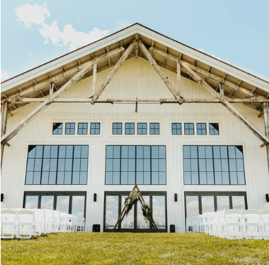Large outdoor wedding venue with a white building, glass doors, chairs arranged outside on the grass, and a clear sky.