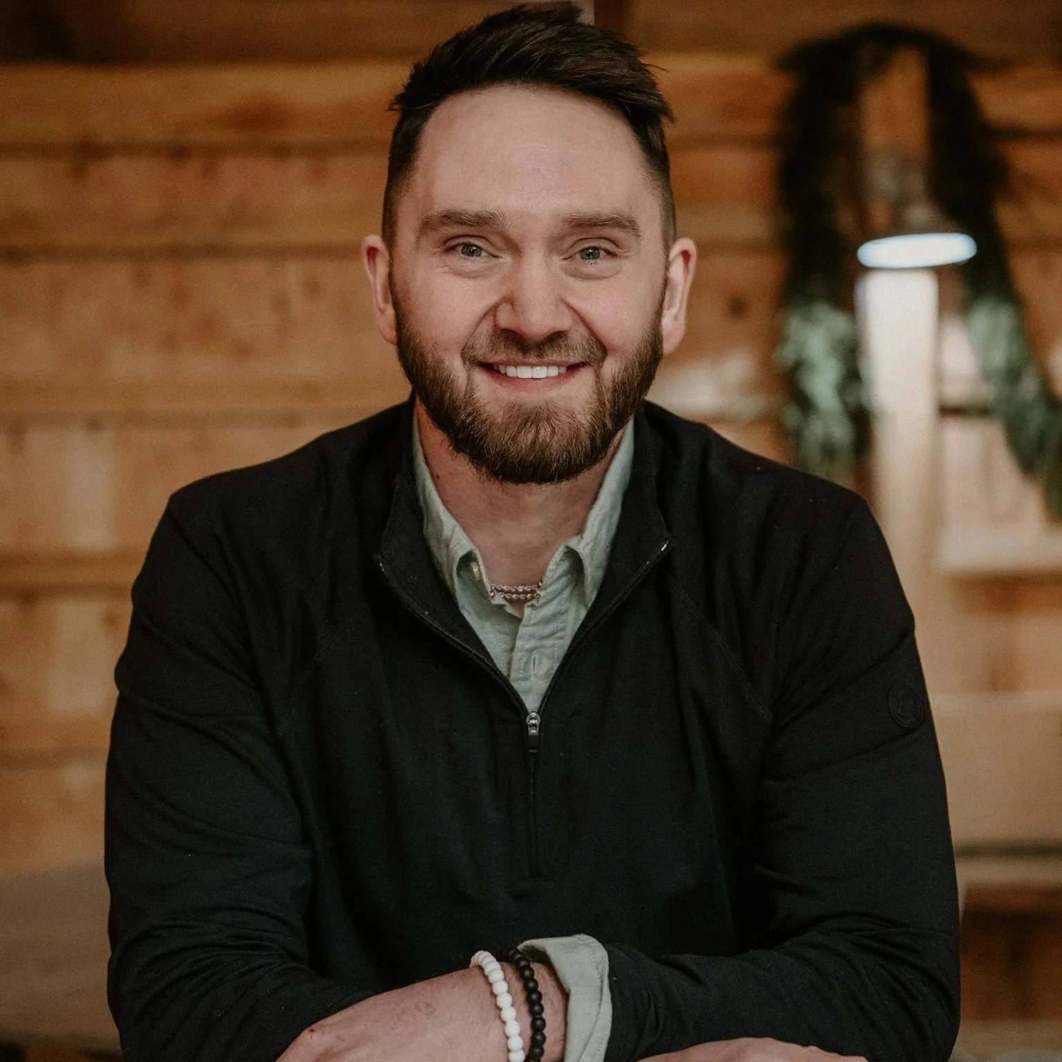 A young man with dark hair, a beard, and light-colored eyes, smiling, sitting behind a bar indoors with a wooden background, wearing a black jacket over a gray shirt, accessorized with jewelry and bracelets.