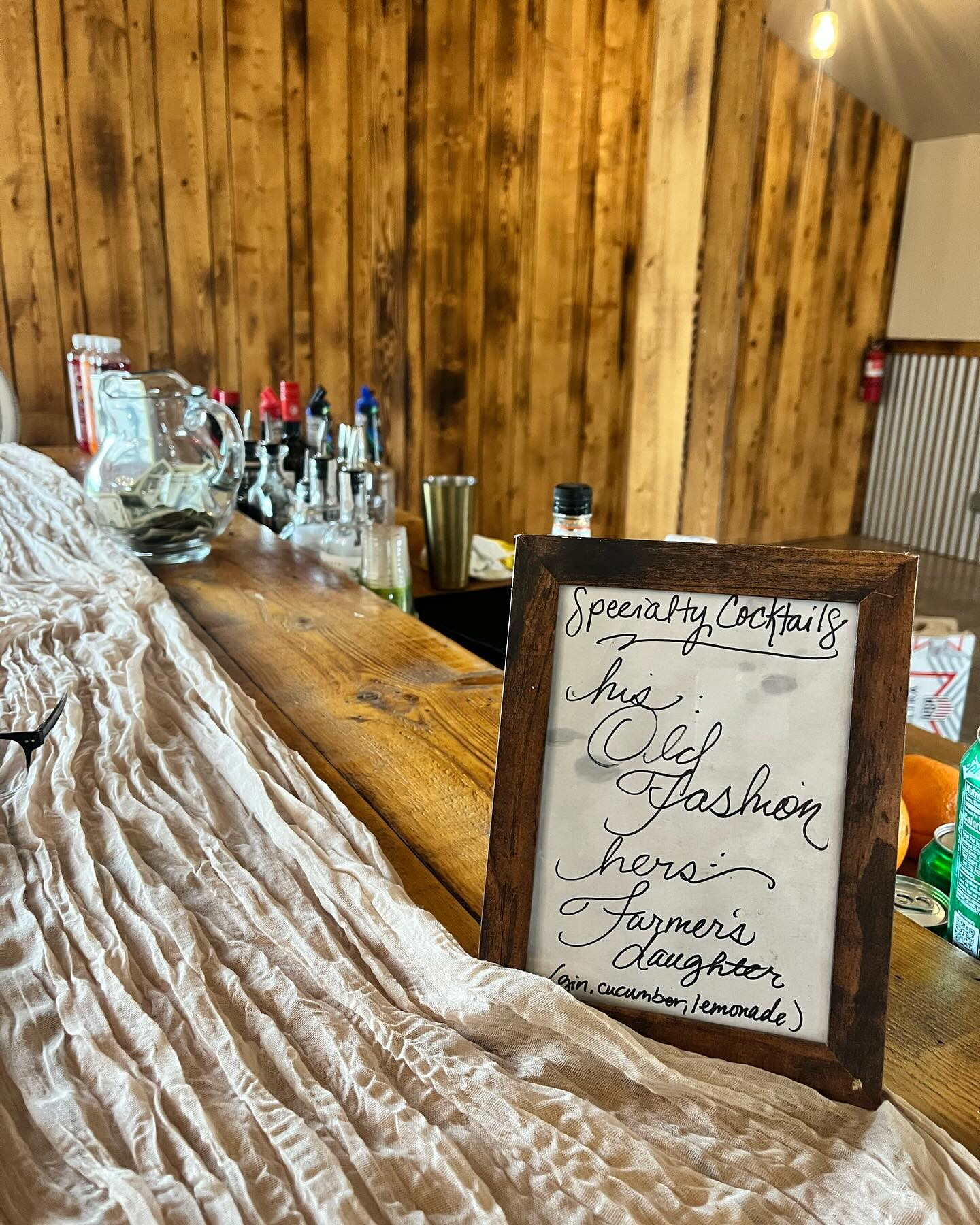 A wooden bar counter with various drink bottles and pitchers. A framed handwritten sign reads 'Specialty Cocktails this Old Fashion cheers Farmers daughter (cucumber, lemonade).' The background has a wood-paneled wall and a fire extinguisher.