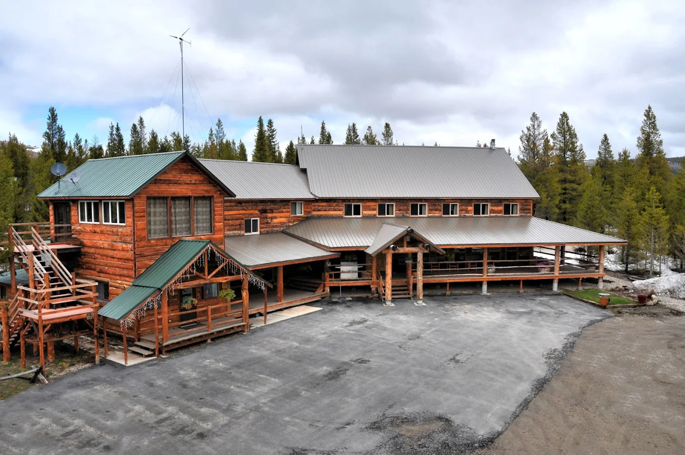A large log cabin with a metal roof, surrounded by trees, with a gravel driveway in front and a small yard on the side under a cloudy sky.