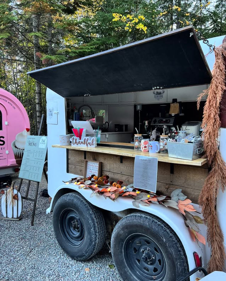 A food truck decorated with fall leaves and a "thankful" sign, serving coffee and food with trees in the background.