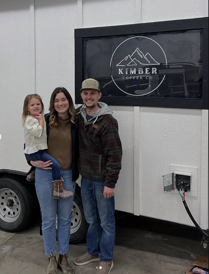 A family of three standing in front of a mobile coffee stand with the logo 'Kimber Coffee Co' featuring mountains inside a circle.