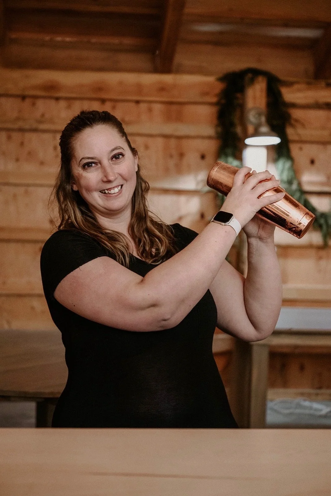 A woman smiling and holding a copper cocktail shaker in a wooden interior setting.