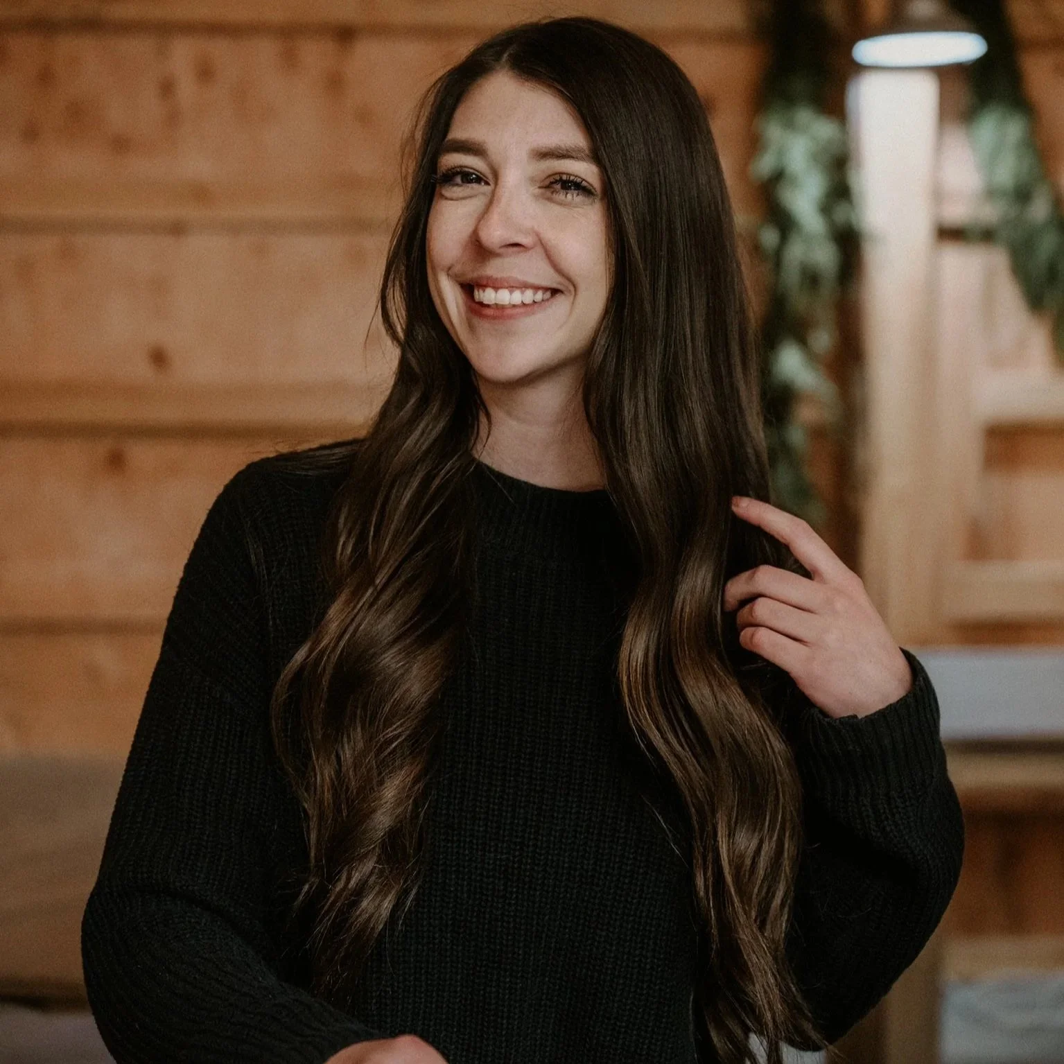 A woman with long brown hair wearing a black sweater, smiling and touching her hair in a cozy wooden interior with greenery in the background.