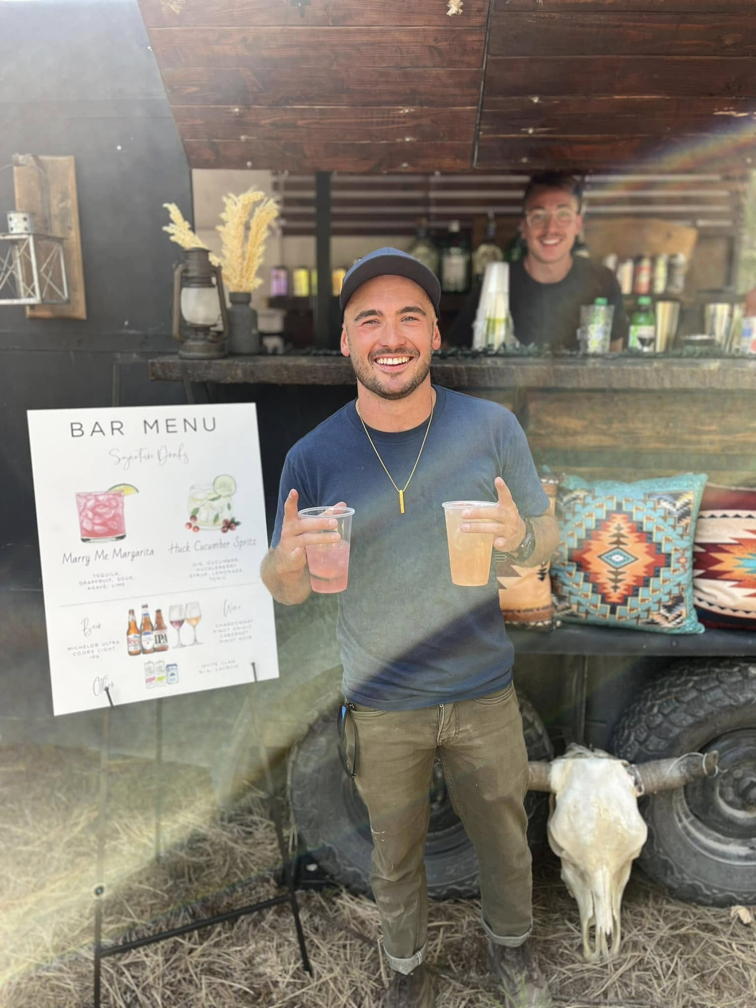 A man wearing a baseball cap, glasses, and casual clothing is smiling and holding two drinks, one pink and one orange. Behind him, there is a bar setup with shelves, bottles, and a sign that reads "Bar Menu" with drink illustrations. Another person w