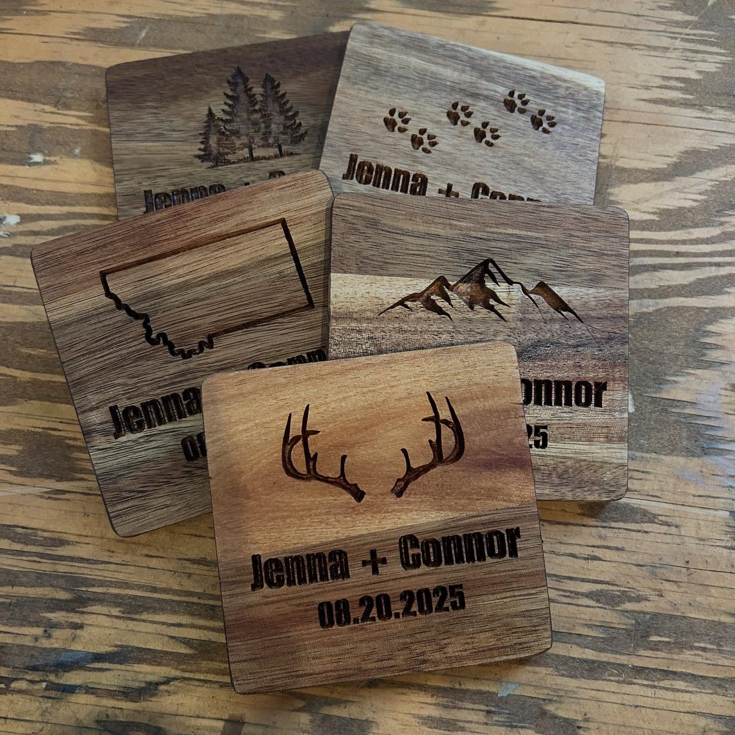 Four wooden engraved wedding date cards with mountain, trees, and paw print designs, featuring names Jenna and Connor and the date 08.20.2025.
