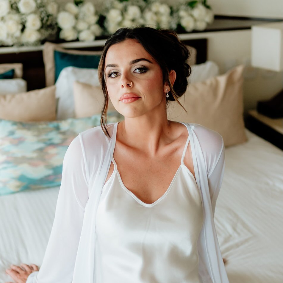 A woman with dark hair and light makeup sitting on a bed with floral and beige pillows in a well-lit room.