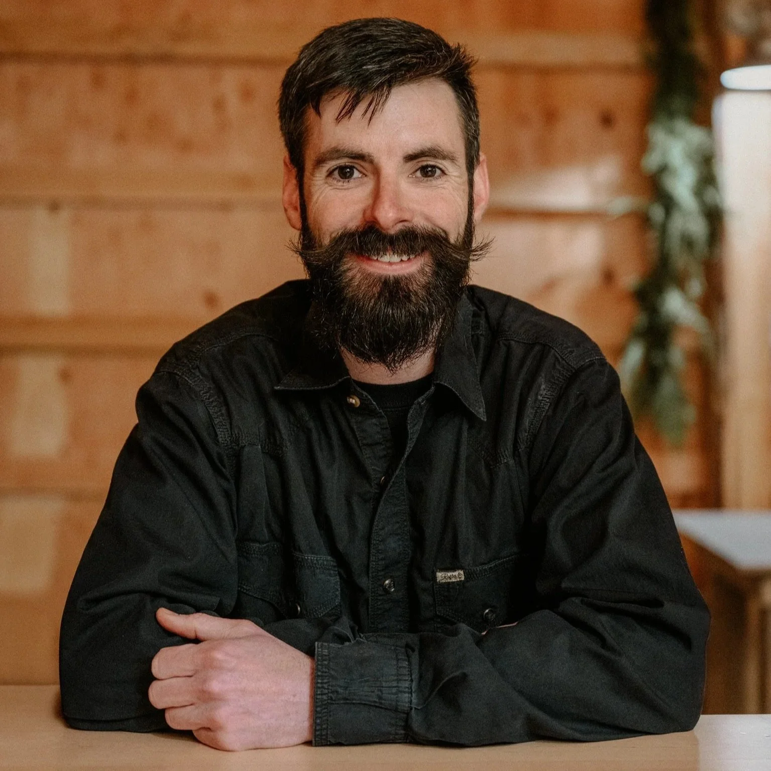 A man with dark hair, a full beard and mustache, wearing a black button-up shirt, sitting at a wooden bar with his arms crossed, smiling at the camera against a wooden wall background.