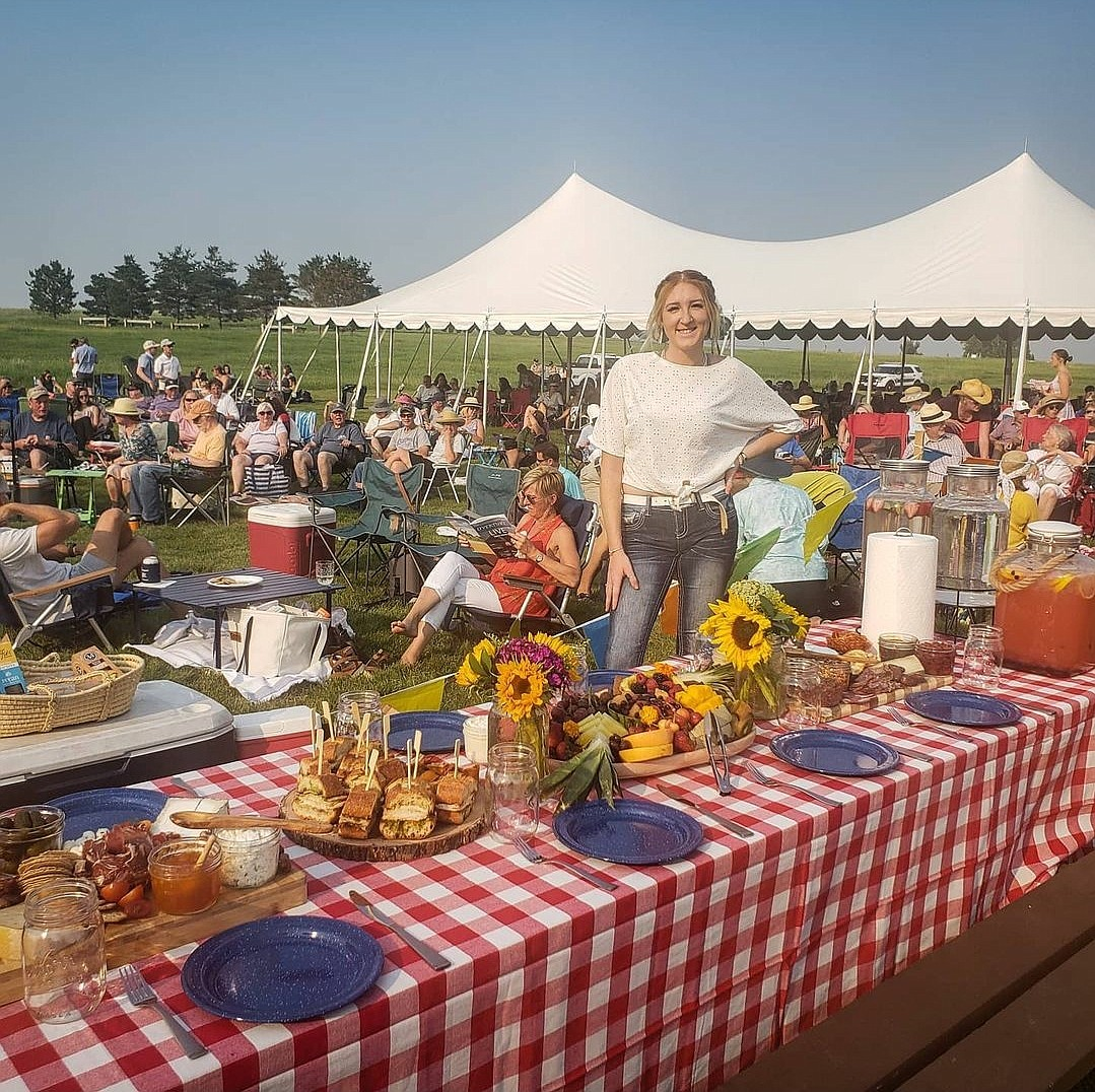 A woman standing behind a picnic table with a red and white checkered tablecloth, filled with food and drinks. In the background, there is a large outdoor gathering with many people sitting under a white event tent on a grassy field.