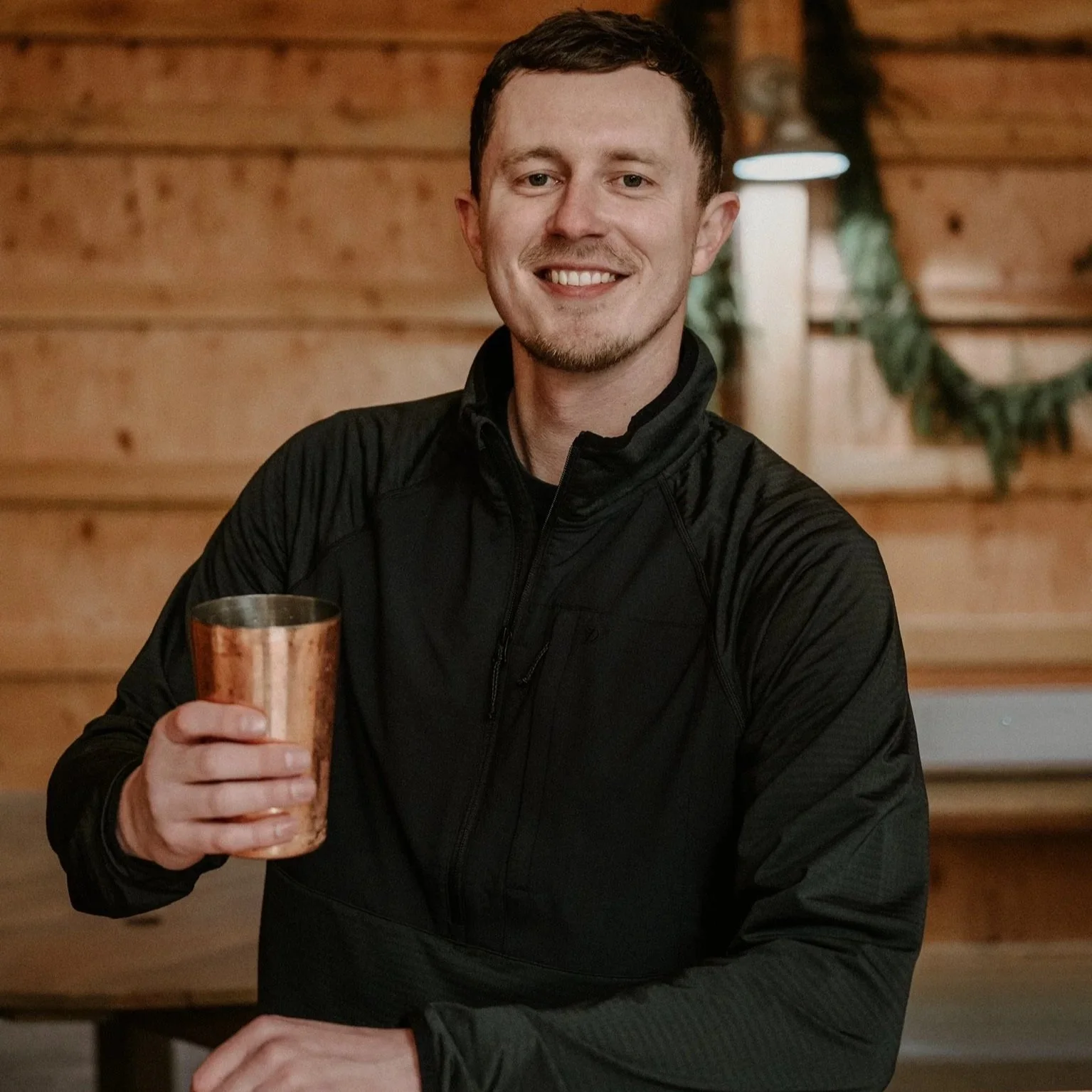 A young man smiling and holding a copper cup, sitting behind a bar in a cozy wooden interior with greenery in the background.