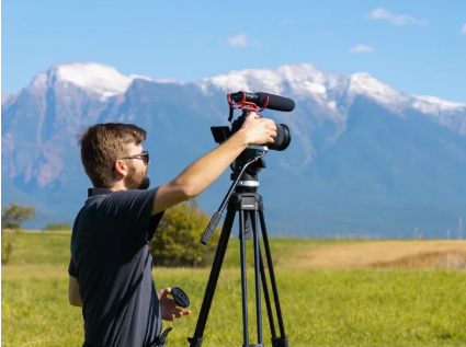 Man with sunglasses operating a telescope on a tripod in a grassy field with mountains in the background.