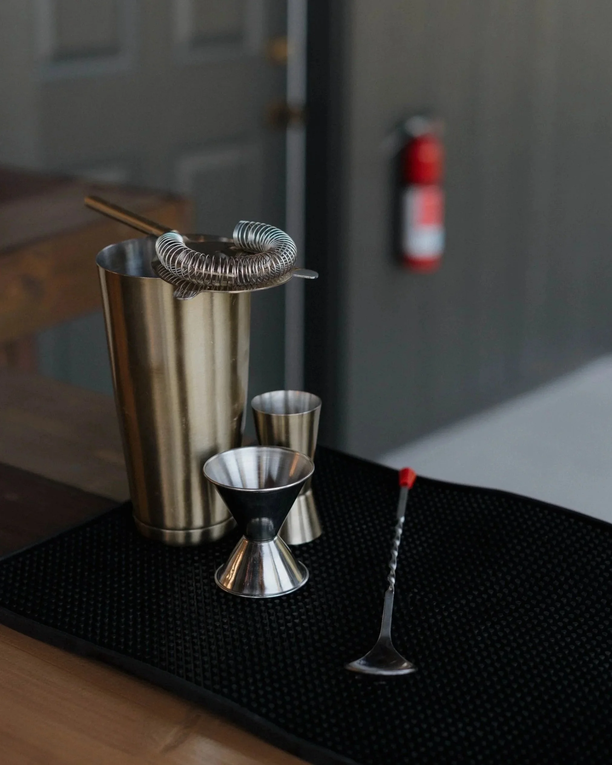 Set of stainless steel cocktail shakers, a cocktail strainer with coiled spring, and a martini glass on a black textured surface.