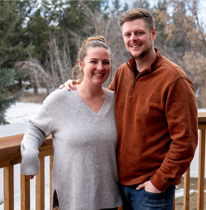 A smiling woman and man standing on a wooden deck outdoors with trees and a snowy landscape in the background.