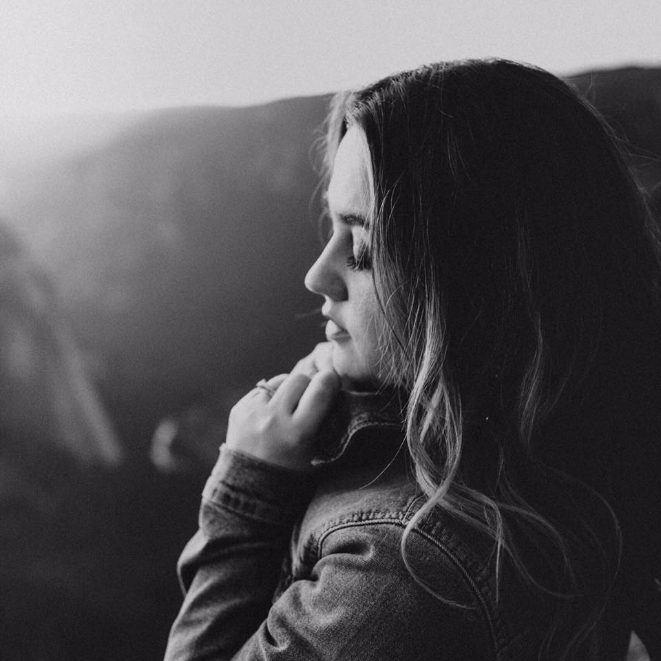 A young woman with wavy hair, wearing a denim jacket, sitting in a contemplative pose with her hand near her chin, looking downward, in black and white.