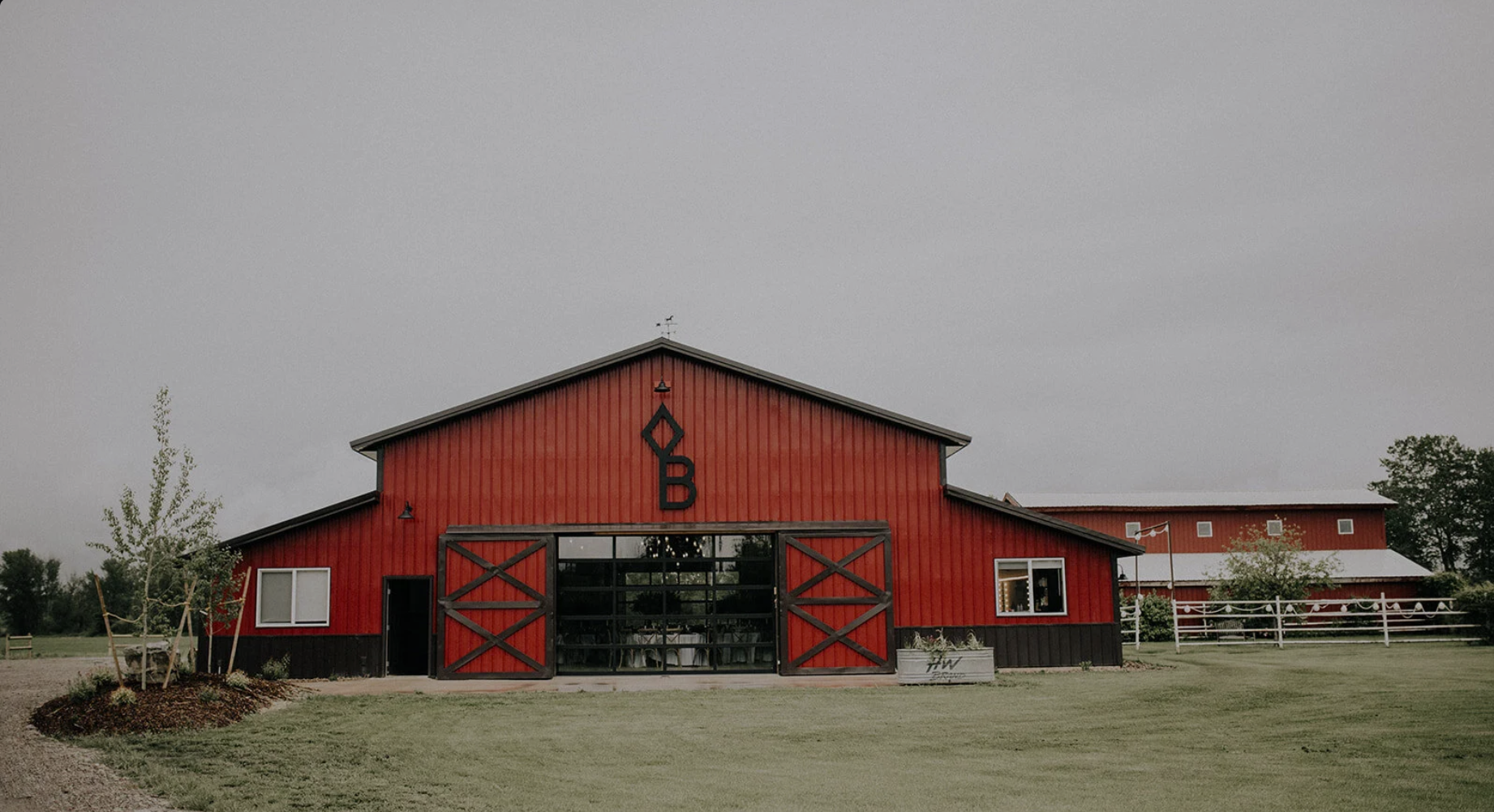 Red barn with black accents and large glass doors, surrounded by a manicured lawn, trees, and a gray sky.