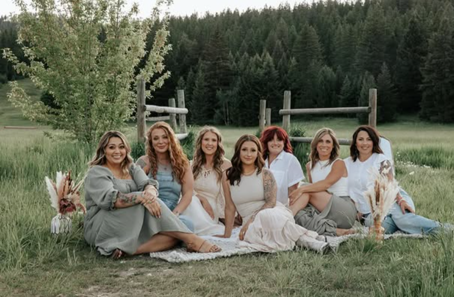 Seven women sitting on a blanket in a field with greenery and trees in the background.