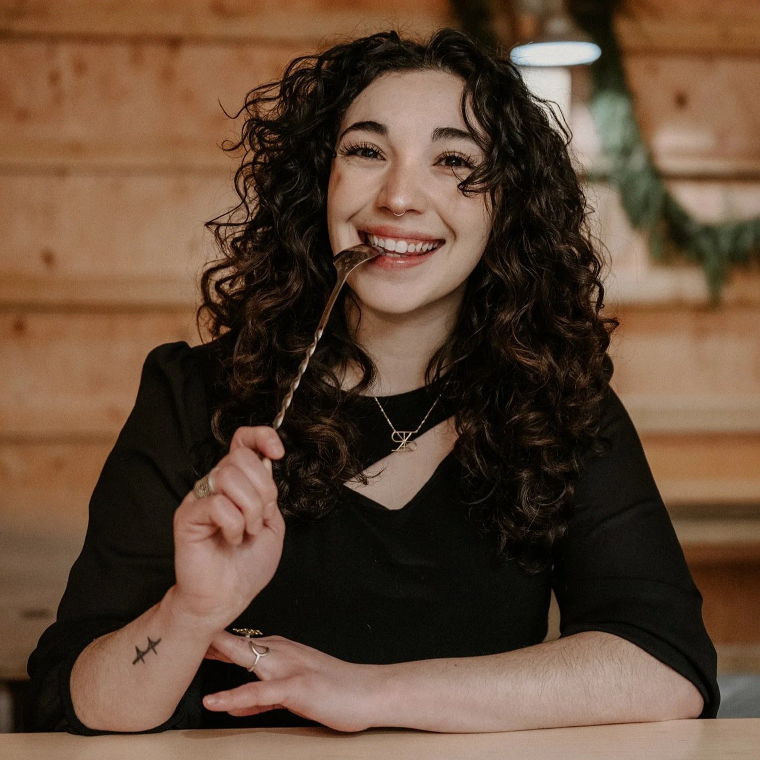 A woman with dark, curly hair smiling and holding mixing spoon in her right hand up to her mouth. She is wearing a black top and sitting at a wooden bar with a wooden background. She has a nose ring, a star tattoo on her right wrist, and a necklace.