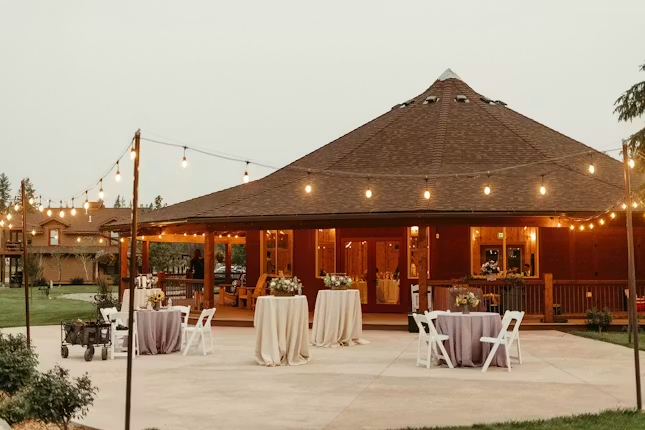 Outdoor event space with round tables draped in white and purple tablecloths, decorated with flower arrangements, under string lights near a pavilion with a brown shingled roof, surrounded by green grass and trees.
