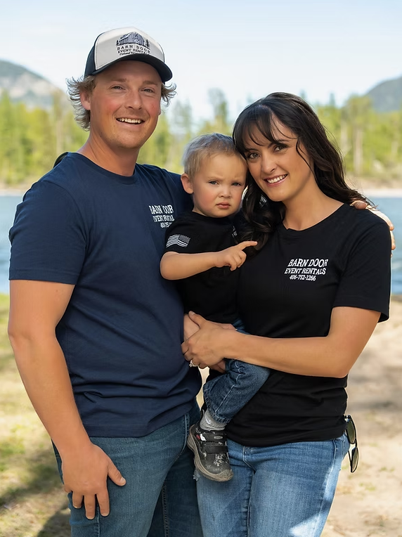 Family of three outdoors with water and trees in the background, smiling at the camera.