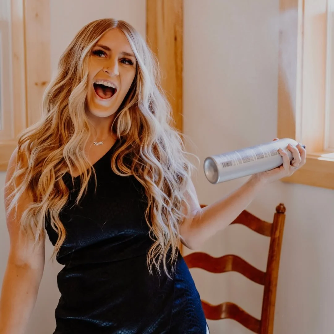 A woman with long, wavy blonde hair, wearing a black dress, joyfully holding a can. She is smiling with her mouth open, standing in a room with wooden accents and a natural light window.