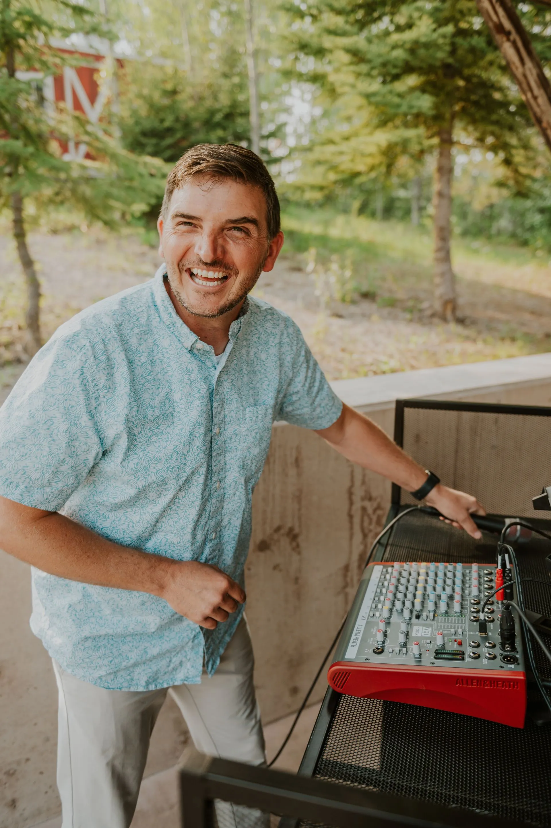 A man in a light floral shirt and beige pants standing outdoors at a music setup with an Allen & Heath audio mixer. He is smiling and looking at the camera. In the background, there are green trees.