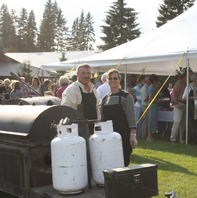Two people stand behind a barbecue grill with propane tanks at an outdoor event, with a crowd under a large white tent and trees in the background.