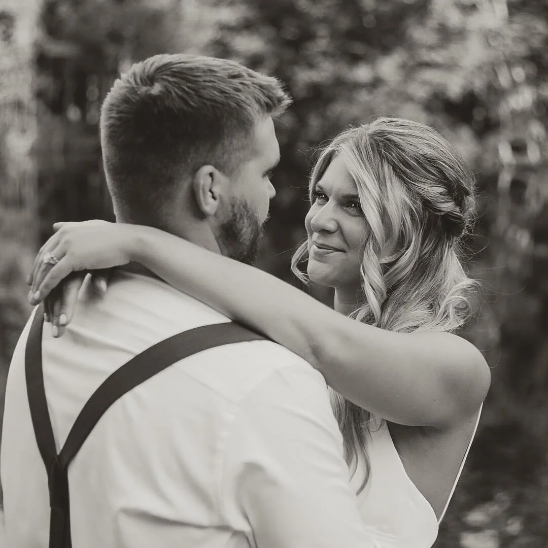 A black and white photo of a couple embracing outdoors, the woman looking into the man's eyes with affection.
