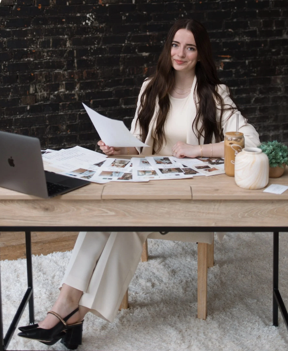 A woman sitting at a wooden desk, holding papers in her hand, with a laptop and photographs spread out on the desk in front of her, against a dark brick wall background.