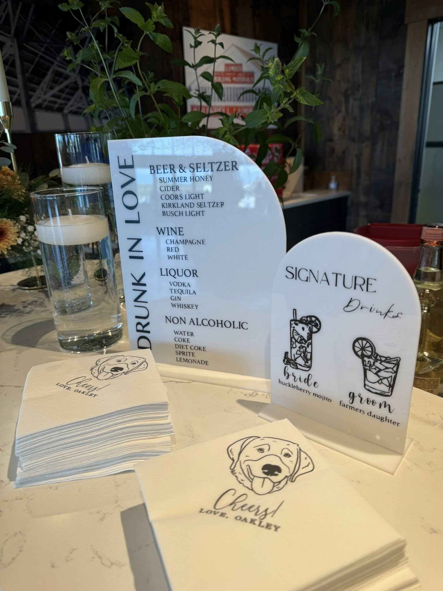 A table with a drink menu, paper napkins with a dog's face illustration, and a card that reads "Cheers!" and "Love, Oakley." The menu lists various alcoholic and non-alcoholic beverages, including beer, wine, and cocktails, with some visible images o