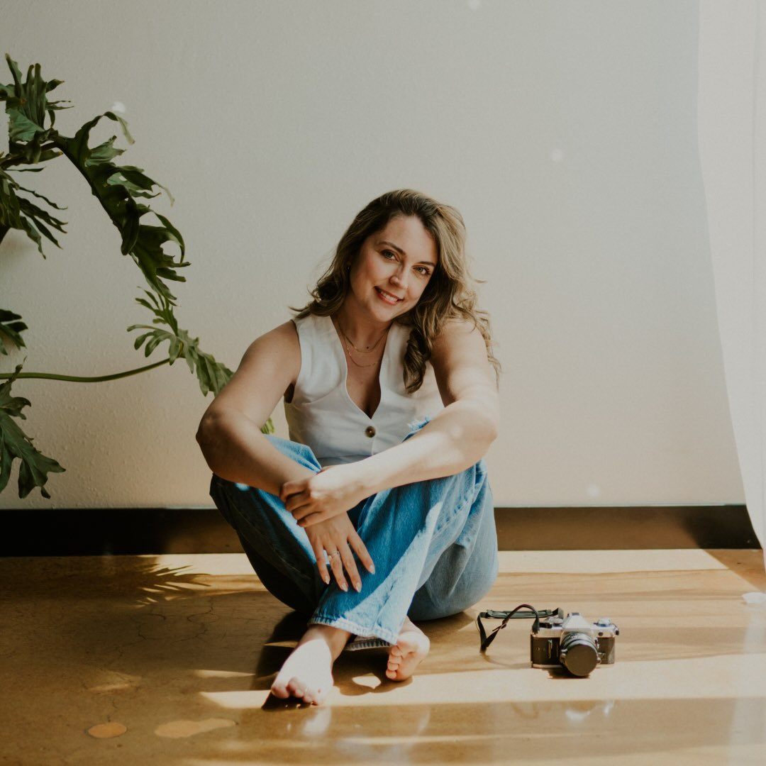 A woman with wavy hair sitting cross-legged on a wooden floor, smiling, wearing a sleeveless top and jeans, with a vintage camera beside her and sunlight streaming in.