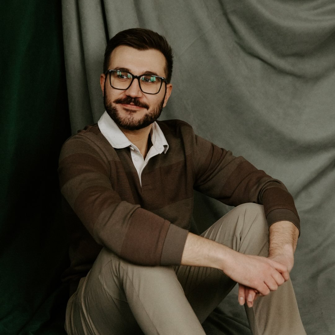 A man with glasses, a beard, and short dark hair, sitting on the floor with crossed legs in front of a gray curtain, wearing a brown sweater over a white collared shirt and beige pants.