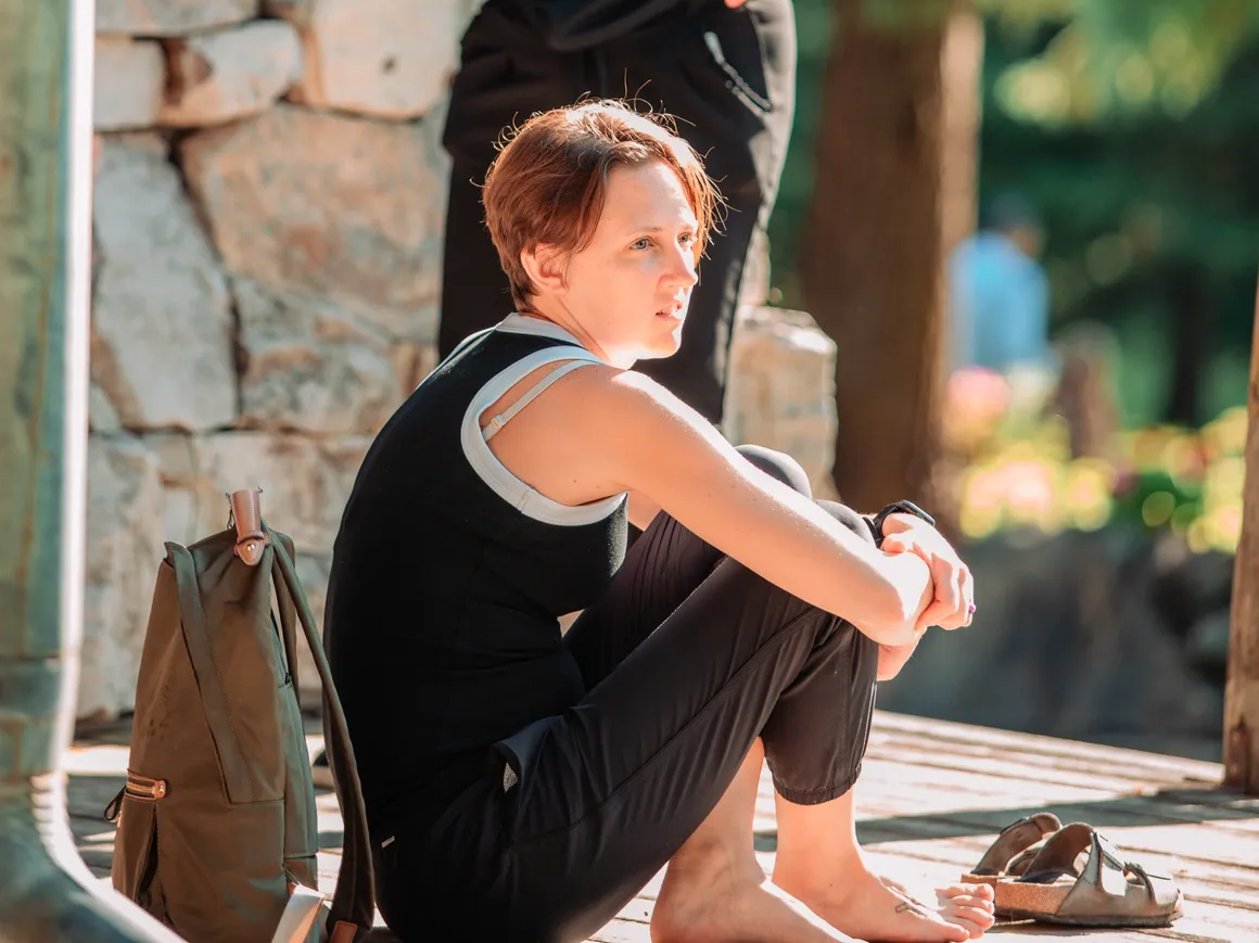 A woman with short reddish hair sitting on a wooden dock, wearing a black tank top and black pants, with sandals nearby and a backpack beside her. She looks pensive or lost in thought.