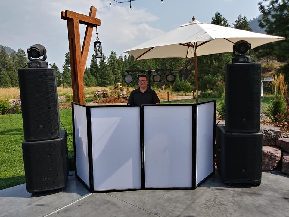 A DJ setup outdoors with large speakers, a white folding screen, a man in black behind the mixer, a large white umbrella, and string lights in a garden with trees and hills in the background.