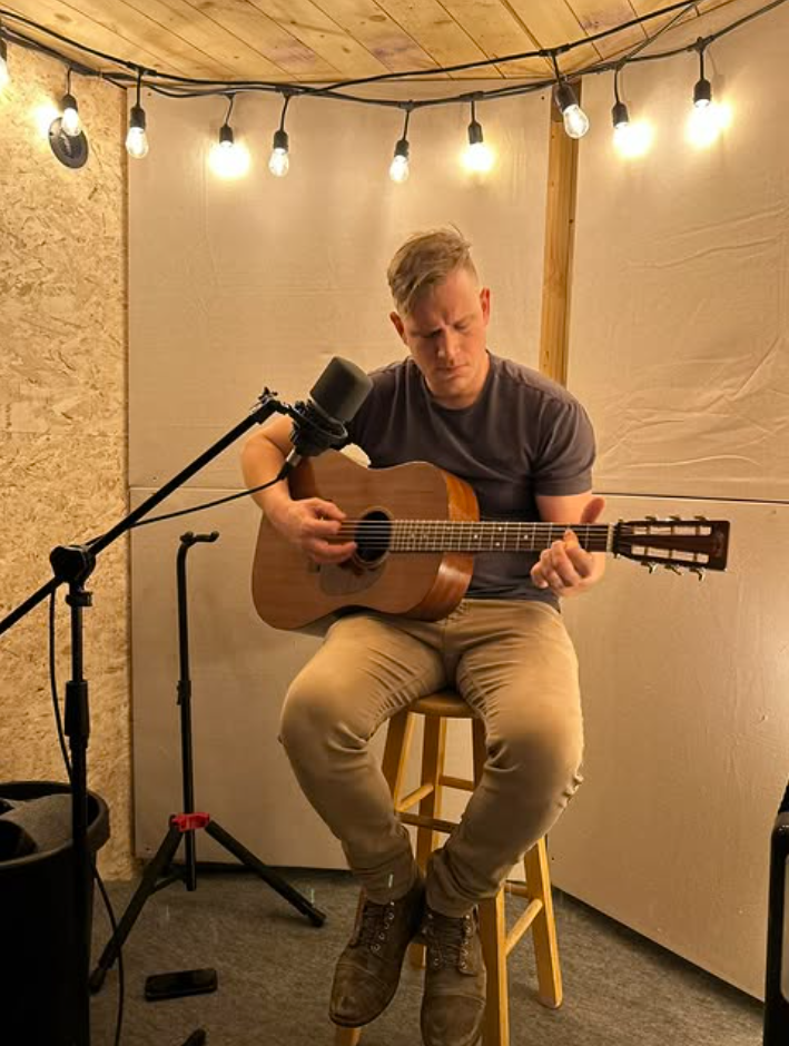 Young man playing acoustic guitar in a small, cozy room with string lights hanging from the ceiling.
