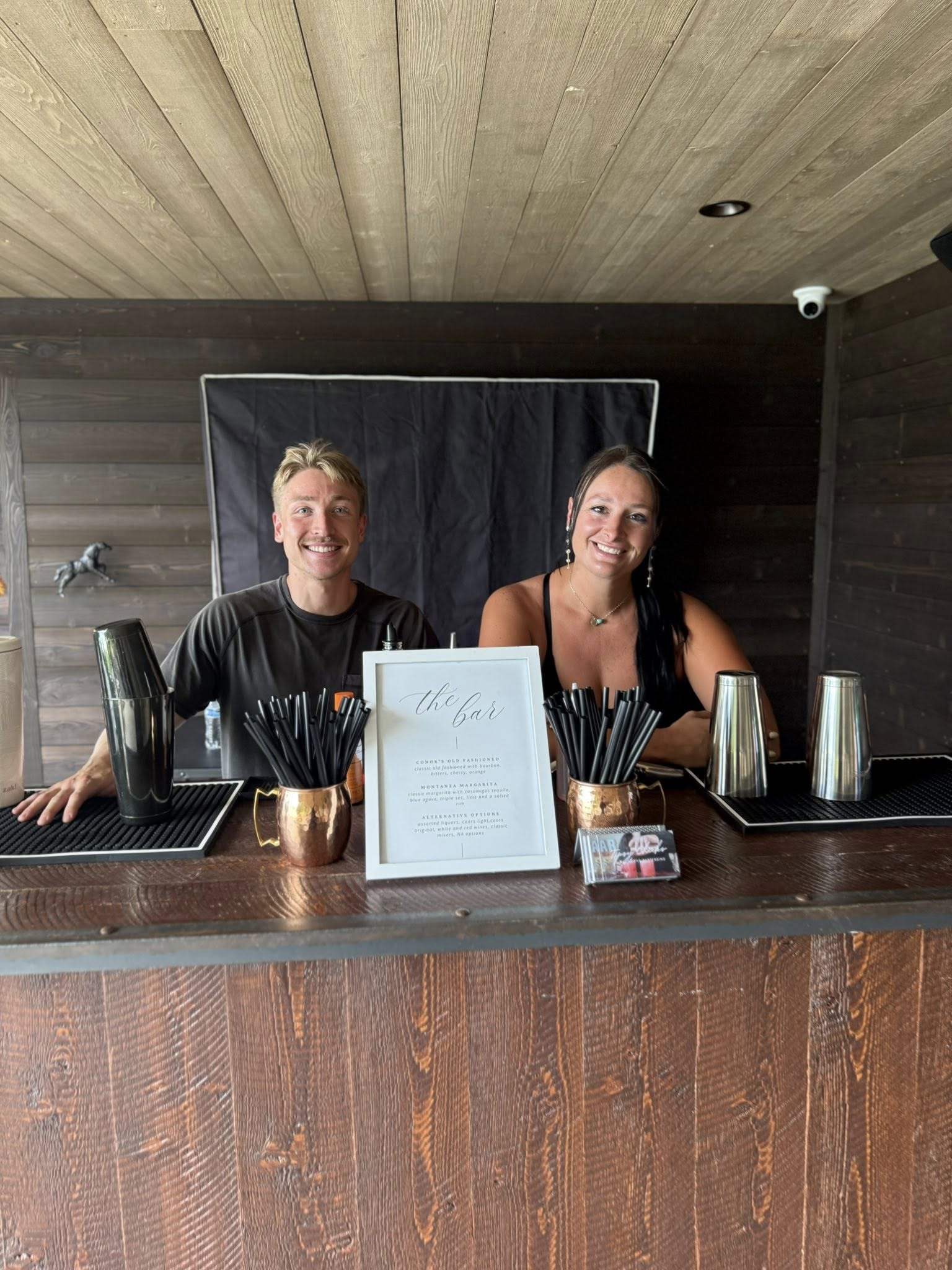 A smiling man and woman sitting behind a bar, with drink straws, a menu, and bar tools on the counter, with dark wood paneling in the background.