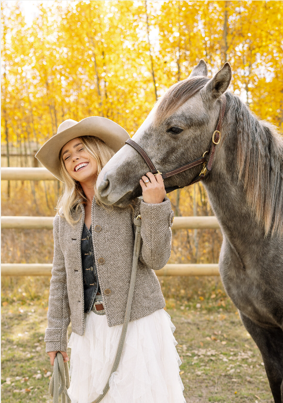 A woman smiling and holding a gray horse's bridle outdoors with yellow autumn trees in the background.