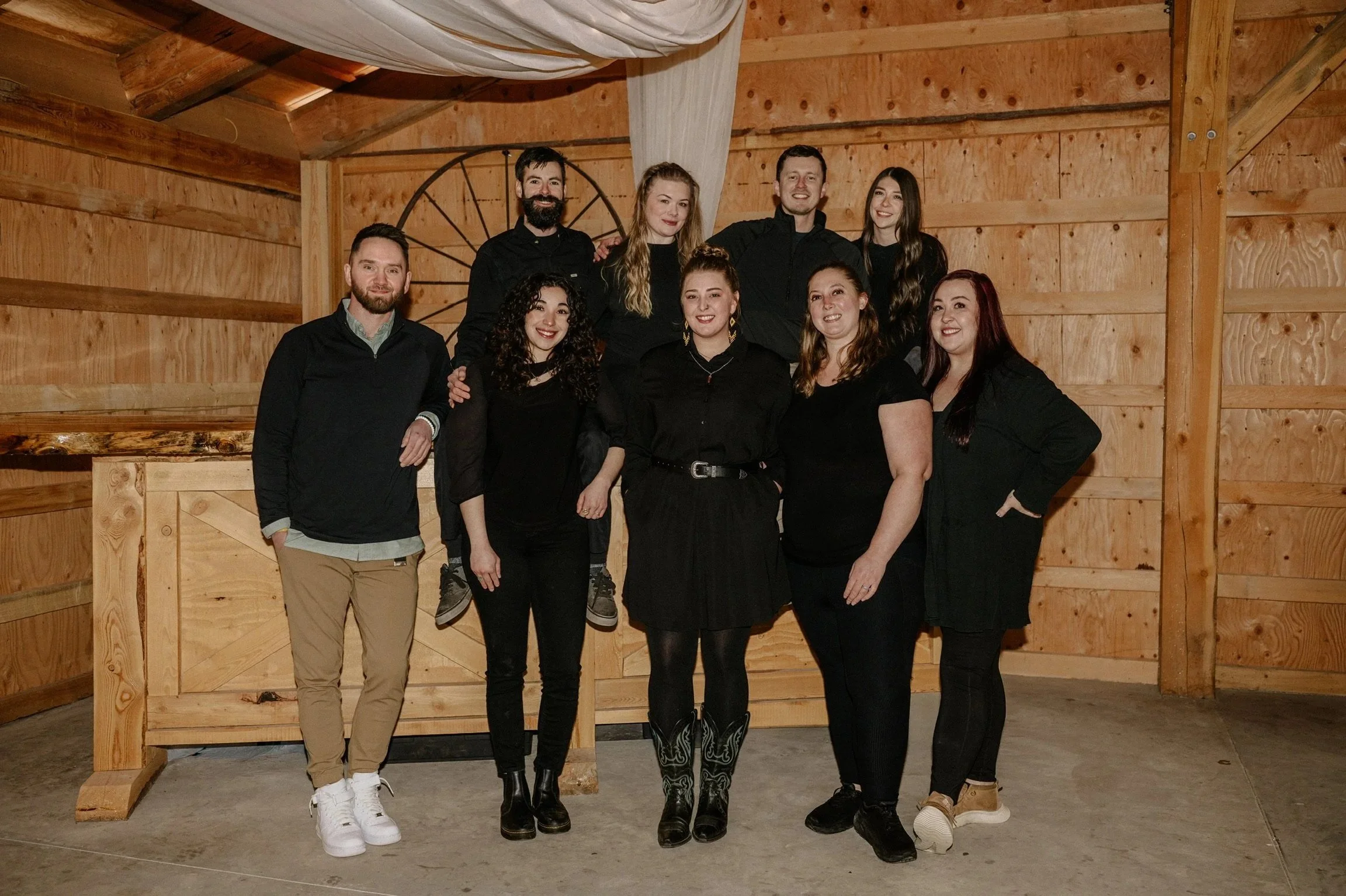 A group of nine bartenders, six women and three men, posing together indoors against a wooden wall with decor, smiling at the camera.