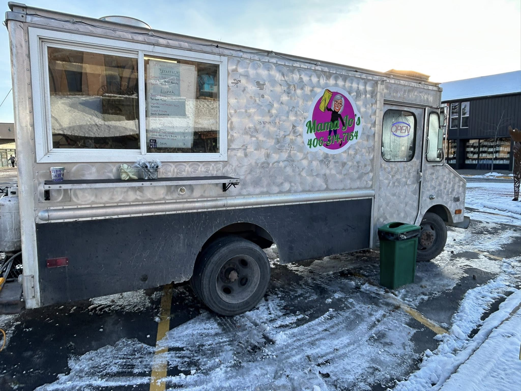 A food truck named Mama Jo's parked in a parking lot on a snowy day. The truck is silver with a large circular logo featuring a cartoon woman with gray hair holding a green bag. The truck has an open window with a menu displayed inside. A small table in front holds a cup and some items. There is a green trash bin nearby, and a building in the background.