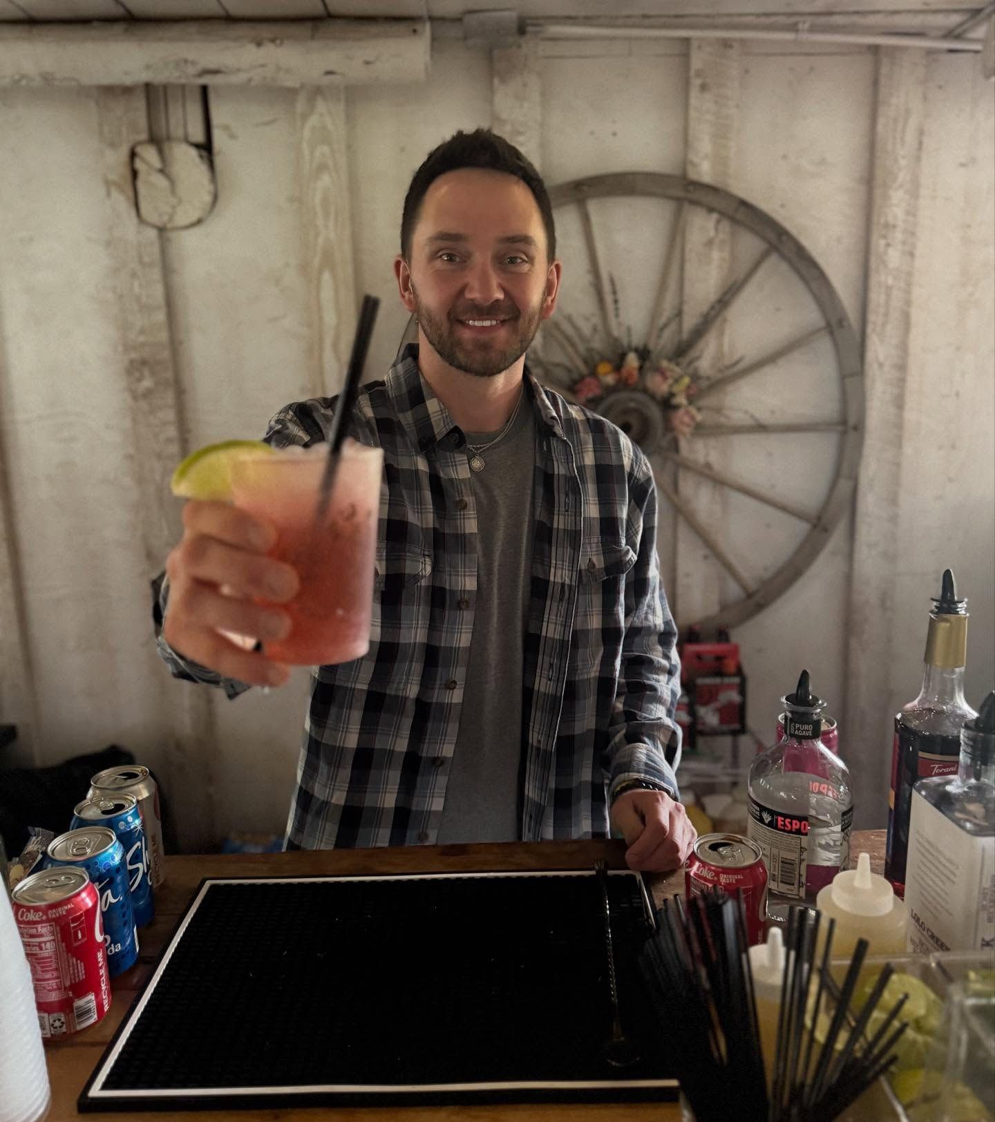 Man smiling and holding a pink cocktail with a lime wedge and black straw, standing behind a bar with soda cans and liquor bottles.