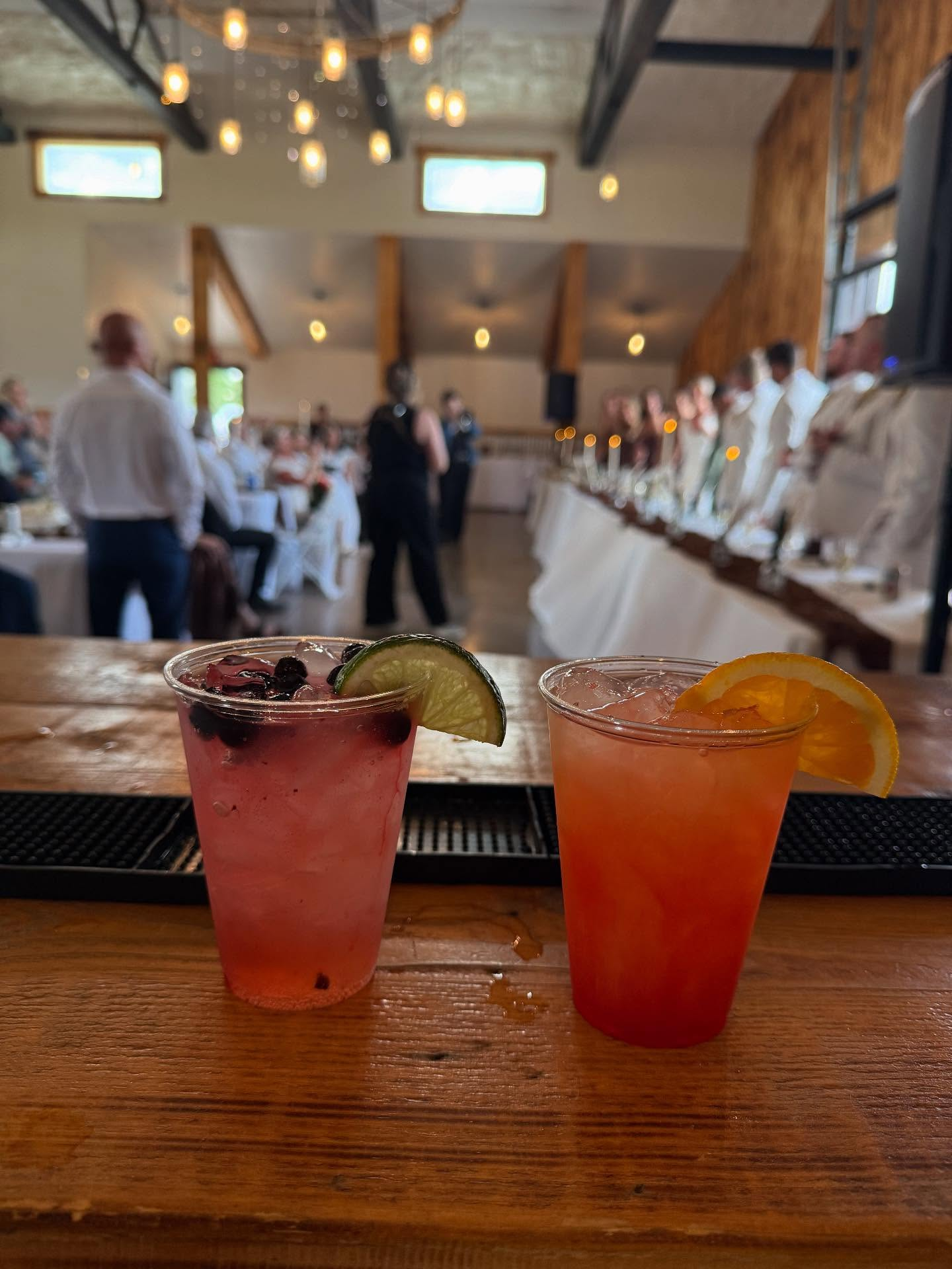 Two colorful cocktails on a wooden bar, garnished with lime, berry, and orange slices, with a blurry background of a dimly lit event venue with people and tables.