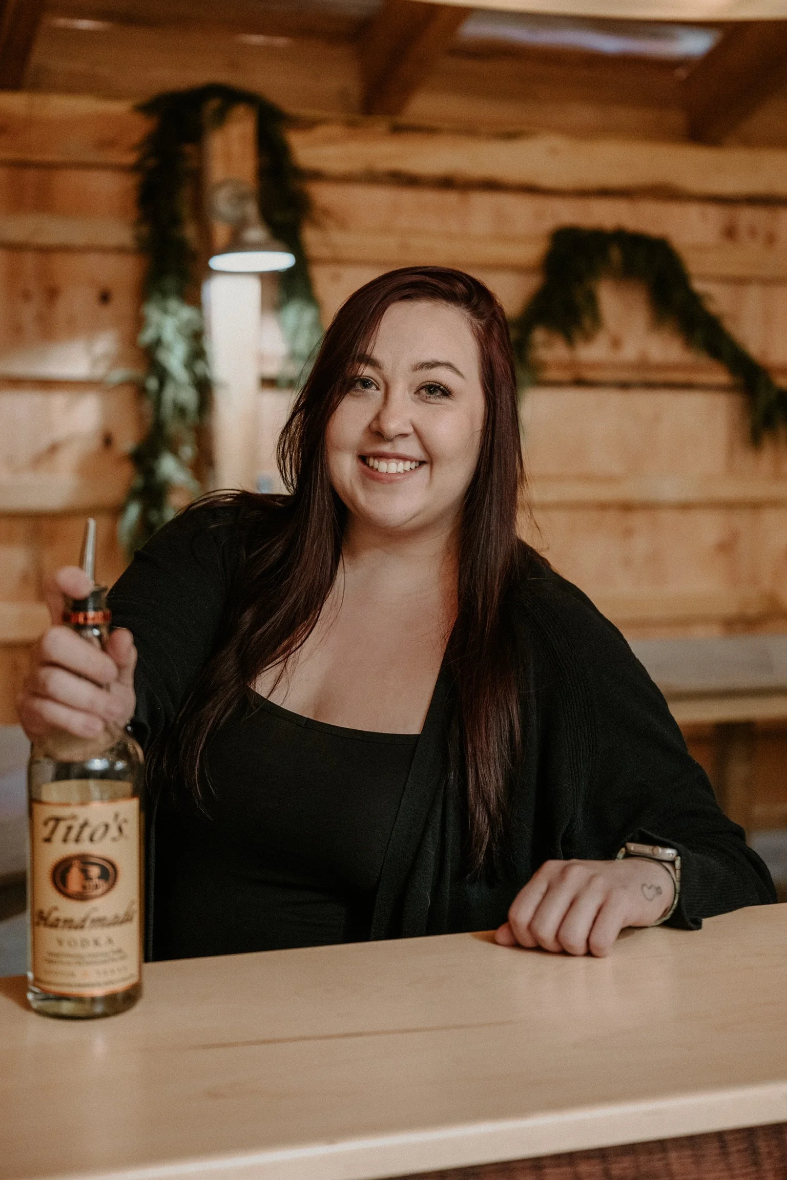 A smiling woman with long dark hair sitting at a wooden bar in a cabin-like wedding venue setting with a bottle of Tito's vodka on the bar in front of her, featuring wooden walls in the background.