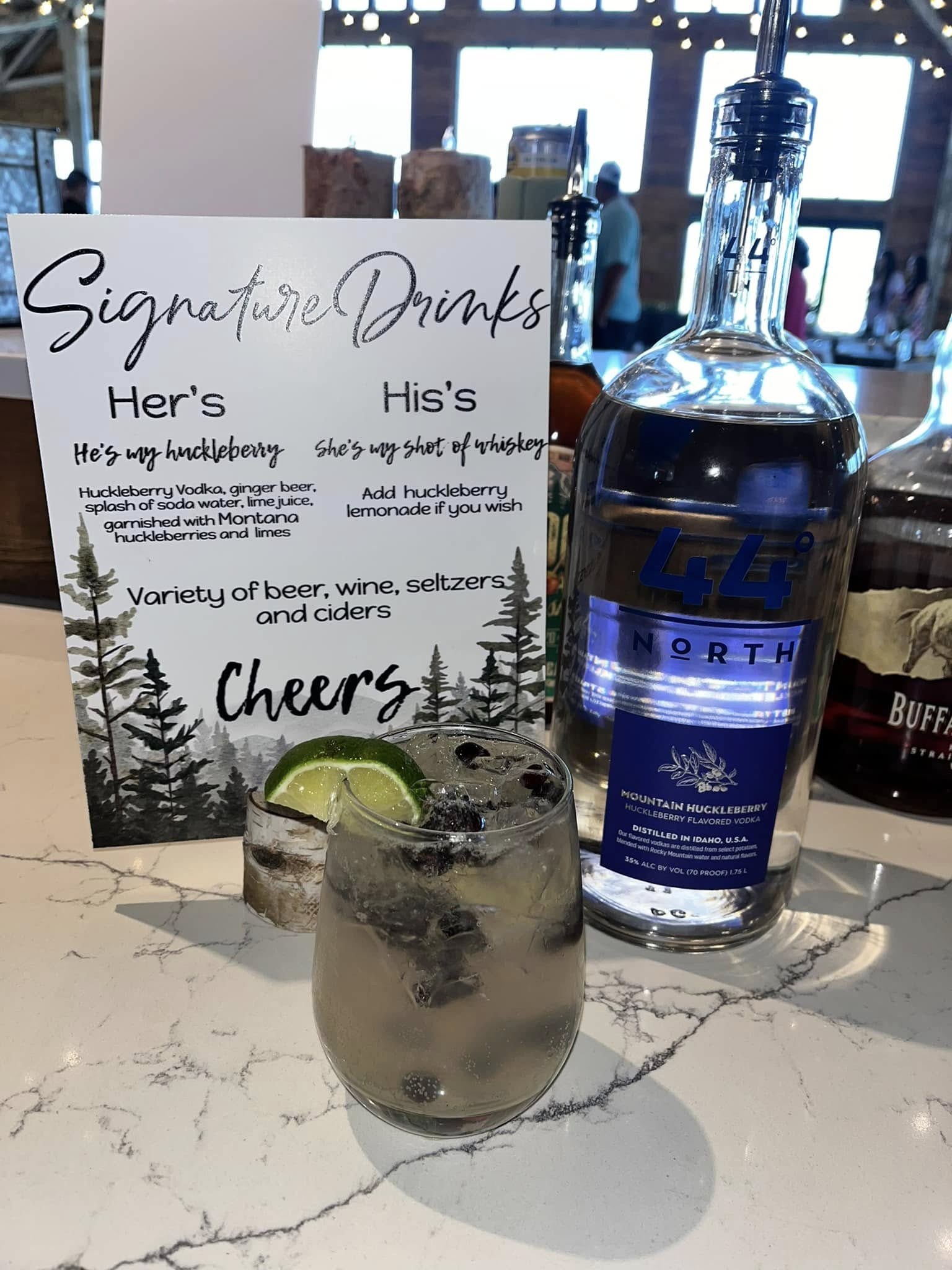 A glass of dark beverage with a lime wedge and blackberries on top, in front of a promotional sign for signature drinks and a bottle of Mountain Huckleberry vodka on a white marble surface.