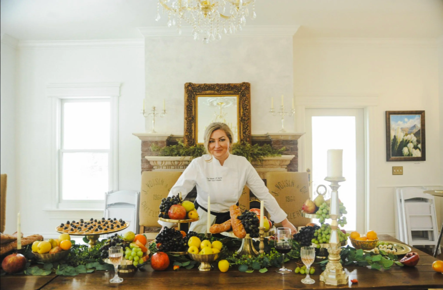 Woman in chef's coat standing behind a festive table with fruits, cheese, and wine, in a bright room with windows, a chandelier, and artwork on the walls.